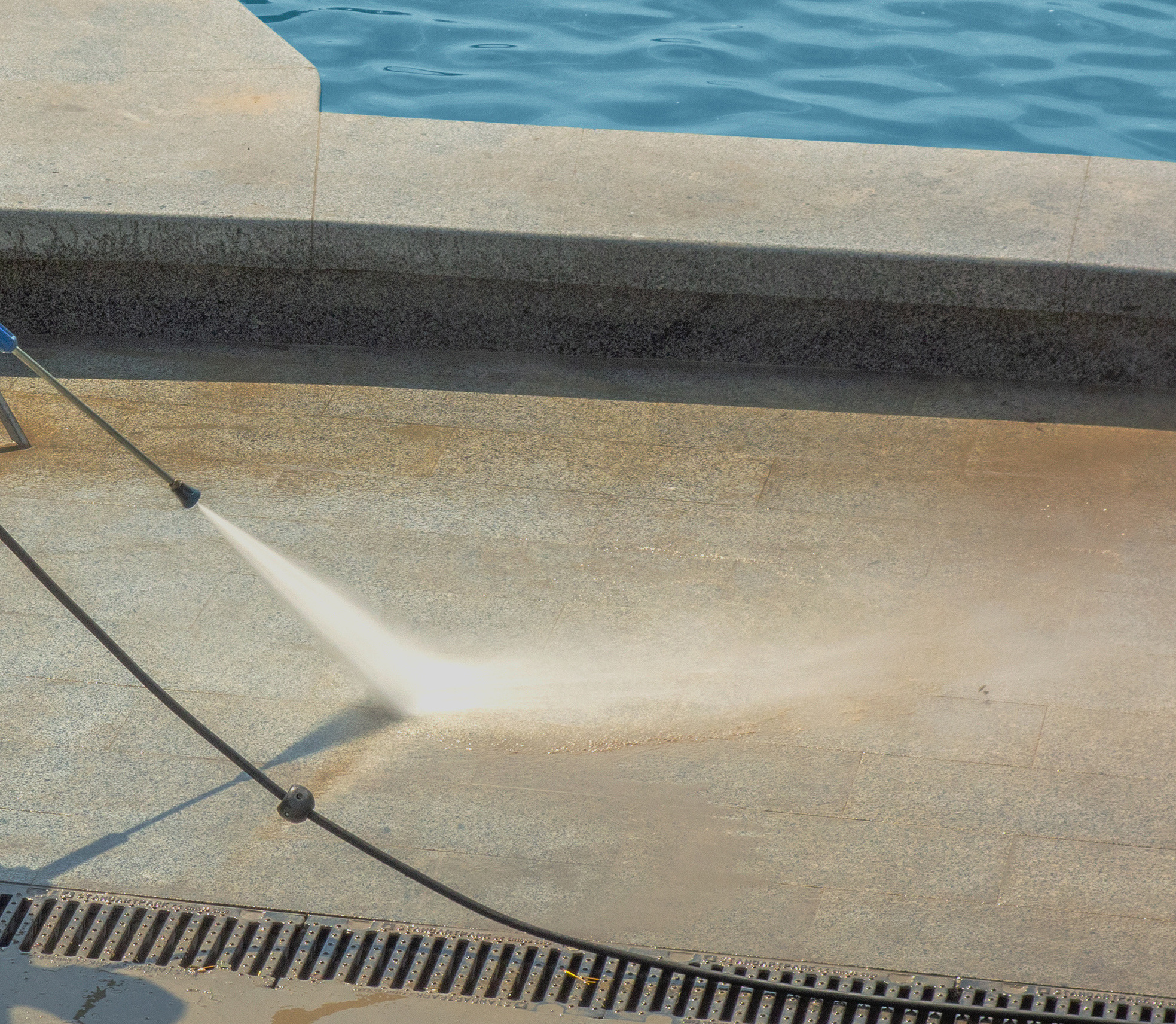 Worker man in uniform washes street or park sidewalk near water pool or fountain. Municipal service of city cleaning process. Guy uses spray equipment